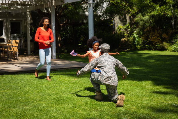 Soldier with family Rear view of a young adult African American male soldier kneeling on the grass while his young daughter runs to greet him and his mixed race wife walks behind her soldiers returning home stock pictures, royalty-free photos & images