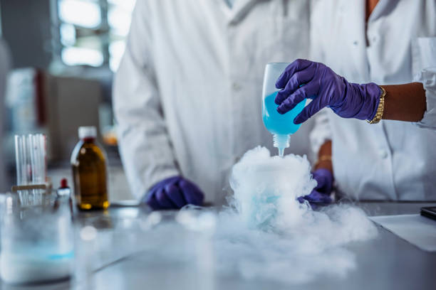 Solution Creating Chemical Reaction in University Laboratory Surface level view of woman wearing white lab coat and purple gloves pouring blue solution into beaker and causing vaporous reaction. chemical reaction stock pictures, royalty-free photos & images
