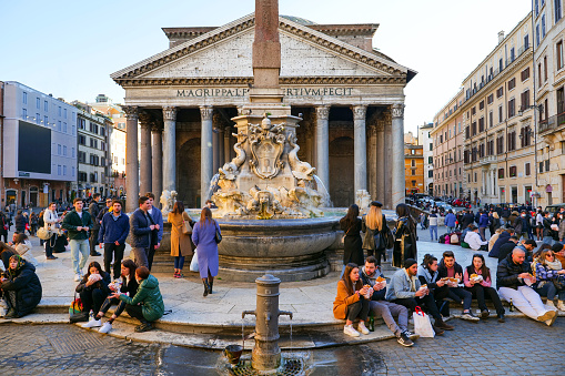 Rome, Italy, January 12 -- Some tourists enjoy a sandwich sitting near the fountain in front of the Pantheon, in the historic heart of the Eternal City. In many historical monuments in Rome and Italy it is normally forbidden to sit down to consume food and drinks. Built in 27 BC by the Consul Marco Vispanio Agrippa for the Emperor Augustus and dedicated to all the Roman divinities, the majestic Pantheon is one of the best preserved Roman structures in the Eternal City and in the world. The fountain in the center of the square (right), with a real Egyptian obelisk, was built by the sculptor Leonardo Sormani in 1575 on a project by the architect Giacomo della Porta. In 1980 the historic center of Rome was declared a World Heritage Site by Unesco. Image in high definition format.