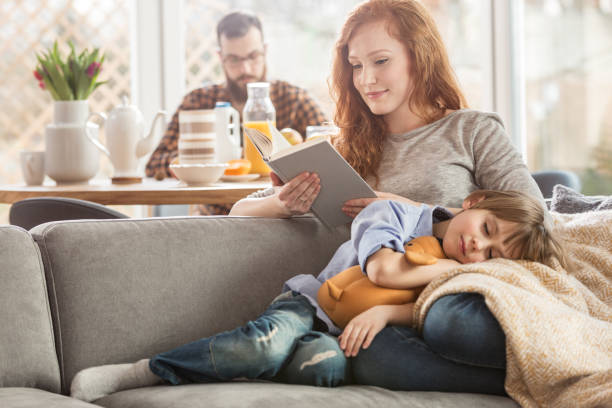 Son lying on mother's laps Sleeping son lying on mother's laps while she is reading a book and father eating in the background comfortable stock pictures, royalty-free photos & images