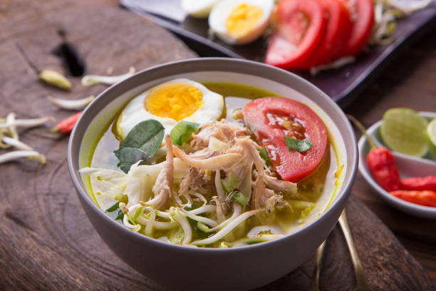 Soto: Indonesian chicken soup with hard boiled eggs, tomato, bean sprout, lemon, cayenne pepper on rustic brown wooden background stock photo