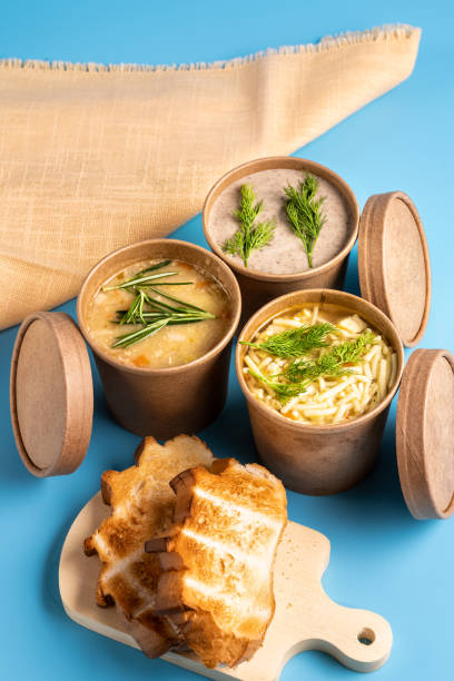 Soups in paper disposable cups for take-out or delivery of food and homemade bread on blue background. stock photo