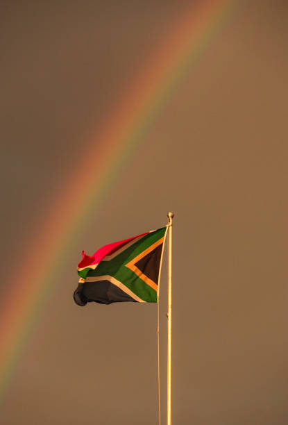A South African flag blowing in a breeze with a rainbow behind it. stock photo