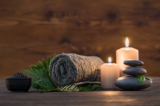 Brown towel on green fern with candles and black hot stone on wooden background. Hot stone massage setting lit by candles. Hot stone therapy for one person with candle light. Beauty spa treatment and relax concept.