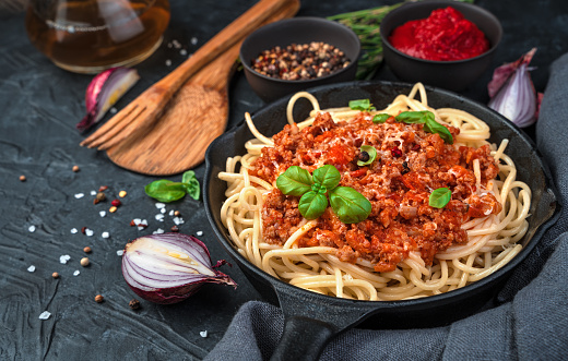 Spaghetti and sauce in a frying pan are sprinkled with cheese and fresh basil on a background of spices and onions on a black concrete background. Side view, horizontal.