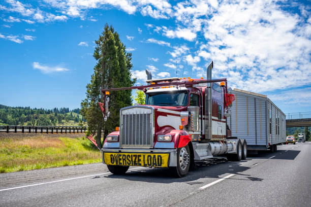 Specially equipped big rig classic red semi truck with oversize load sign transporting manufactory house on step down semi trailer running on the road with escort car stock photo