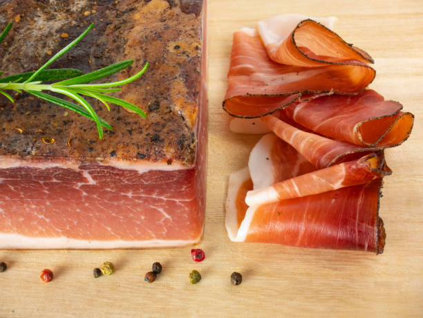 Speck with slices on the cutting board. Typical South Tyrolean smoked bacon. Sliced raw ham. Dry cured meat. Traditional cold cuts, Italian speck with rosemary and pepper. Still life food. stock photo