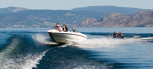 Speed boat with two people on a tube behind it on a lake Action photo of power boat pulling tubers on Okanagan Lake motorboat stock pictures, royalty-free photos & images