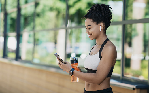 Sports blog and social networks to chat with followers. Cheerful millennial african american female in sportswear and wireless headphones holding bottle of water, typing on smartphone and relax