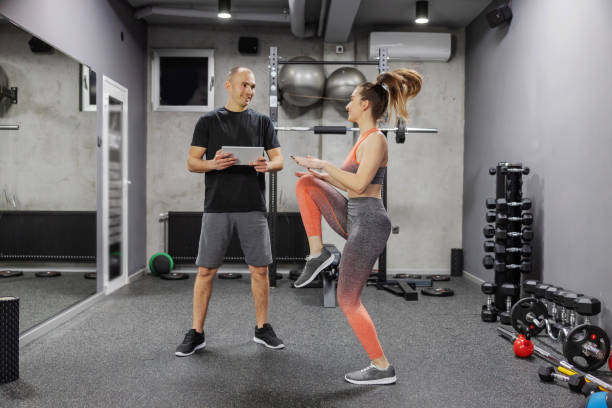Sports training performance and progress measurement. A woman in sportswear does a high skip indoor gym while the trainer observes her training and enters data into a digital tablet stock photo