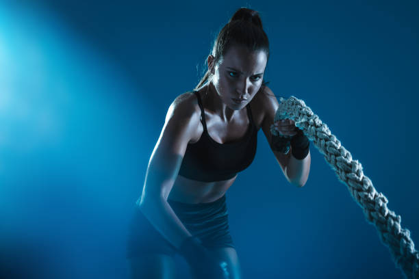 Sportswoman exercising with battling ropes Athlete moving the ropes in wave motion as part of fat burning workout. Sportswoman exercising with battling ropes on blue background. battle stock pictures, royalty-free photos & images