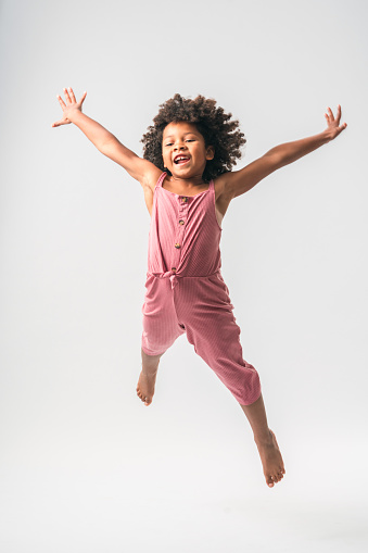 Cute black-haired, curly girl in pink jumpsuit, jumping off the ground in front of a white studio background, arms stretched away from the body and looking away.