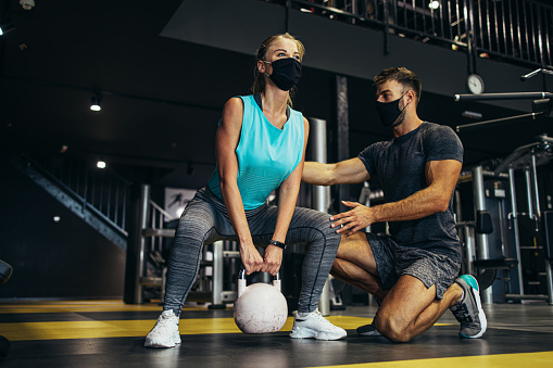 Young fit and attractive woman at body workout in modern gym together with her personal fitness instructor or coach. They keeping distance and wearing protective face masks. Coronavirus world pandemic and sport theme.