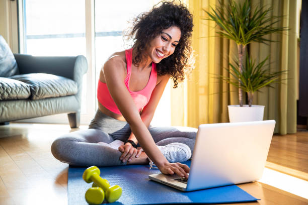 Sporty woman in sportswear is sitting on the floor with dumbbells using a pc laptop in the living room - Young girl training fitness at home - Sport and recreation concept. stock photo