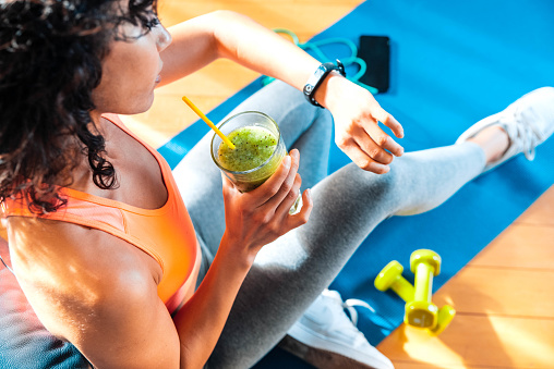 Sporty woman in sportswear training at home drinking fresh smoothie - Fit female athlete using smart watch to monitor her performance - Sport, food and technology concept.