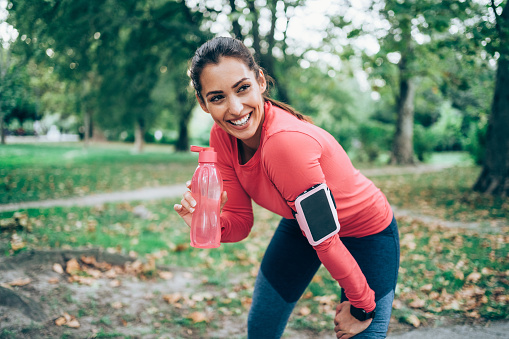 Sporty woman drinking water from bottle after exercising outdoors. Beautiful young woman sipping water from pink bottle after workout at park.