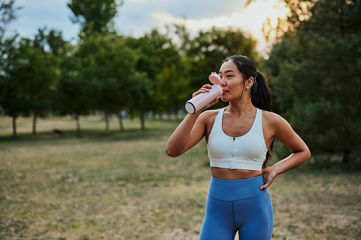 Asian sporty woman resting after workout in the park and drinking water from the battle