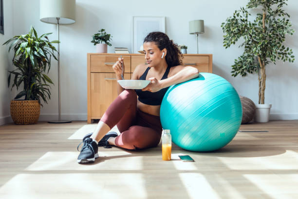 Sporty young woman eating healthy while listening to music sitting on the floor at home. stock photo