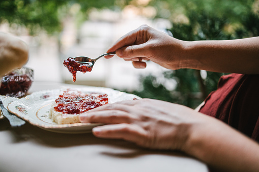 Close-up of woman's hands spreading raspberry jam on a piece of bread. Retro tableware set and natural environment