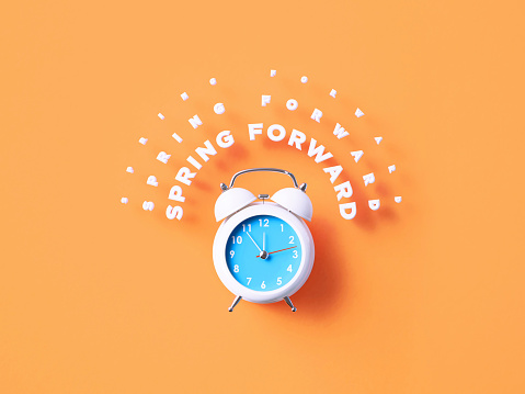 Spring Forward message sitting over white alarm clock on orange background. Horizontal composition with copy space.