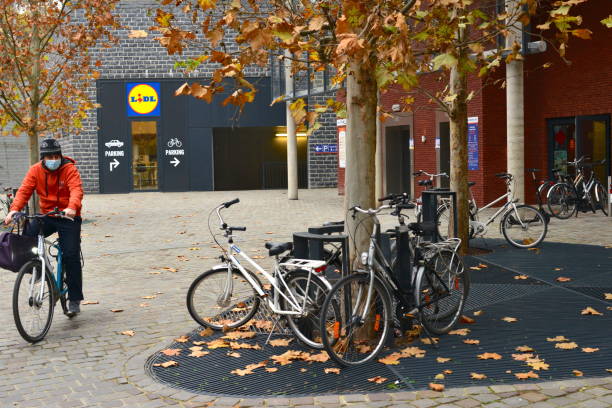 Square Lidl German chain supermarket in Leuven Belgium. Leuven, Vlaams-Brabant, Belgium - November 27, 2020: Lidl Belgium food supermarket chain in autumn. Facade Lidl and Kruidvat. Customer riding on a bike. Parked bicycles.Tree leaf fall. lidl stock pictures, royalty-free photos & images
