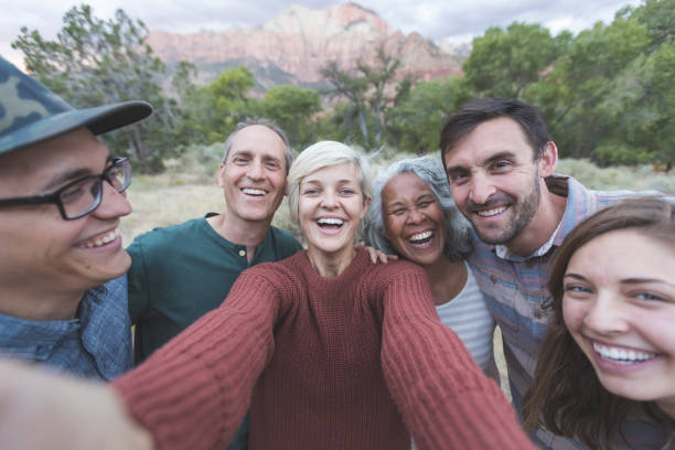 Squeeze in for a group photo! A mixed race senior couple pose with their adult children and friends for a group photo outdoors. A young woman at the center of the frame is holding out the camera and is taking a selfie of the group. Everyone is smiling and laughing. They are standing in a field embracing one another. A forest and mountains are in the background. It is near sunset. The group is on vacation together. friendship photos stock pictures, royalty-free photos & images
