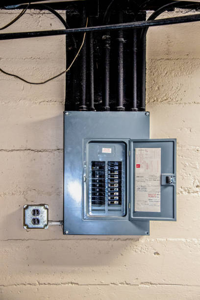 Stainless steel electrical circuit box open with circuits named by hand in basement on rough concrete block wall with conduit and ceiling and other pipes overhead painted black with dust on them stock photo