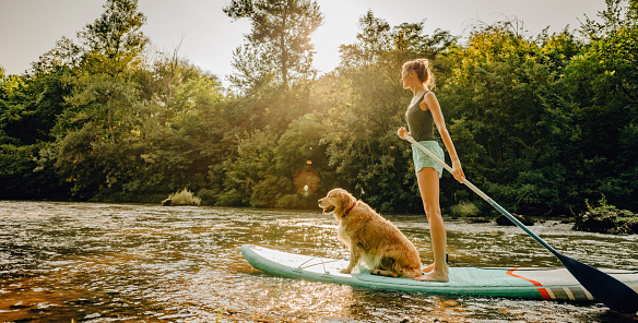 Photo of a young woman and her dog stand up paddling on the river; enjoying the beautiful, warm summer afternoon, far from the hustle of the city.