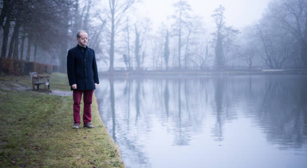 Standing at the very edge of the empty lake, a middle aged man alone in misty forest, lonely and abandoned in gloomy atmospheric mood stock photo