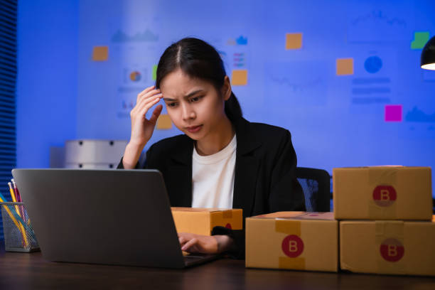 Startup small business concept, young woman owner hands touch on the forehead have a headache because of stress, and checking online order on digital laptop with packing on the box at home office. stock photo