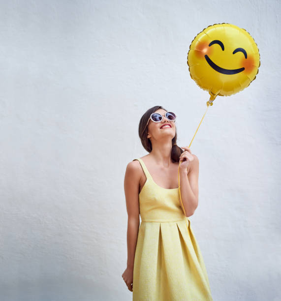 Stay happy! Studio shot of a cheerful young woman holding a smiling emoticon balloon while standing against a grey background big smile emoji stock pictures, royalty-free photos & images