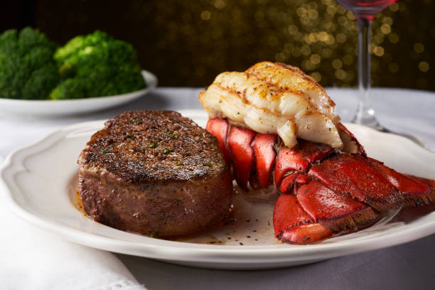 Steak and Lobster on a White Plate and Table Cloth A juicy steak next to a succulent lobster in a white fancy restaurant. There is a plate of blurry broccoli in the background, and this delicious meal is paired with a red wine in a wine glass shown to the right. There is light in the background bokeh into octagons. ornate photos stock pictures, royalty-free photos & images