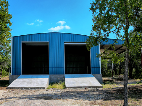 Large blue metal building on country side with open gates on blue  sky background.