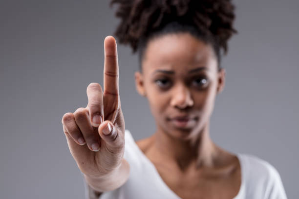Stern focused young woman holding up her index finger Stern focused young woman holding up her index finger as though emphasising a point, calling a halt, or identifying herself on a grey studio background assertiveness stock pictures, royalty-free photos & images