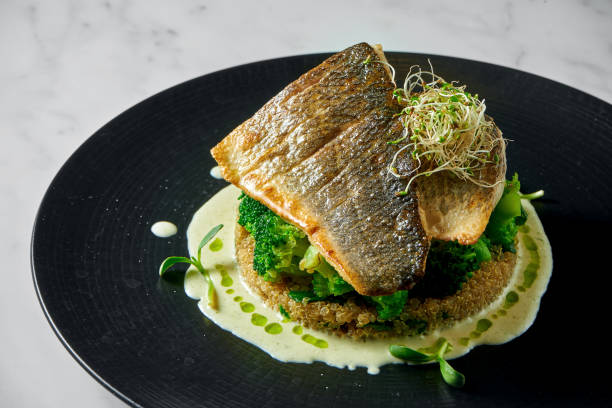 Stir-fried sea bass fillet with quinoa and broccoli garnish with creamy sauce, served in a black plate on a marble background. Appetizing, diet food stock photo