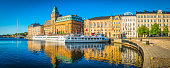 Warm summer sunlight illuminating the ferries on the harbour waterfront reflecting the iconic facades of the luxury hotels and townhouses that line the shore of Nybroviken harbour, Stockholm, Sweden's vibrant capital city.