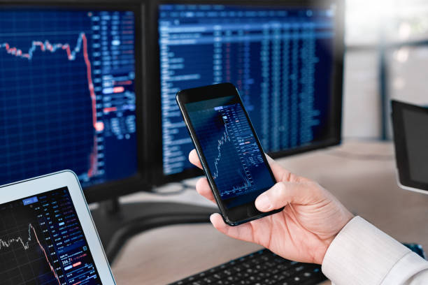 Stocks and Funds. Trader sitting at office in front of monitors with data using app on smartphone monitoring price changes close-up Man trader sitting at desk at office in front of monitors with stocks data holding smartphone browsing application monitoring price changes on candle bar online using digitla devices close-up bitcoin photos stock pictures, royalty-free photos & images