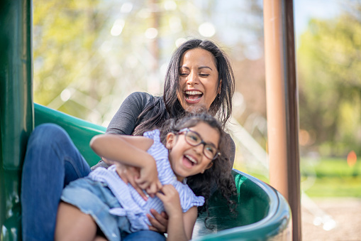 An adorable pair of mother and daughter of Hispanic ethnicity is going down the slide at a playground. They are both happy and smiling.