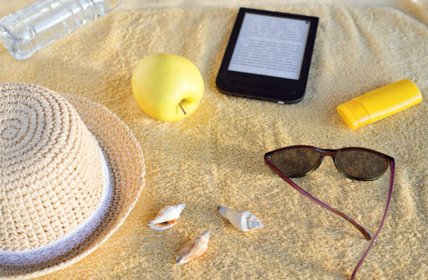 Straw hat, seashells, sunburn cream, sunglasses, apple, bottle with water and e-book reader on beige beach towel. Soft evening sunlight. stock photo