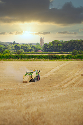Straw or hay Baler machine pulled by a tractor, discharging a compacted straw bale from the rear in a field of recently harvested barley on the outskirts of Cirencester in the Cotswolds