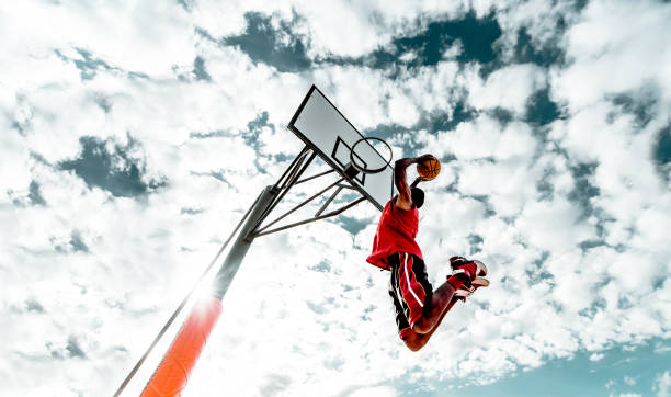 Street basketball player making a powerful slam dunk on the court - Athletic male training outdoor on a cloudy sky background - Sport and competition concept stock photo