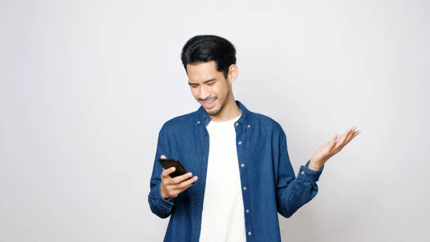 Stress asian man struggle with mobile phone, Frustrated asia male holding smartphone while standing over grey isolated background, People struggle with technology stock photo