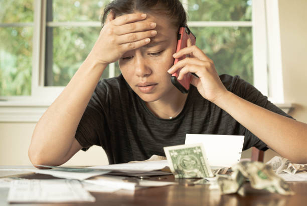 A stressed woman with no money looking at her credit card bills and monthly payments. Financial crisis debt. Poor person sitting at the table at home looking over all her bills and credit card fees with 1 dollar bills and coins lying around, stressed worried look on her face. struggle stock pictures, royalty-free photos & images