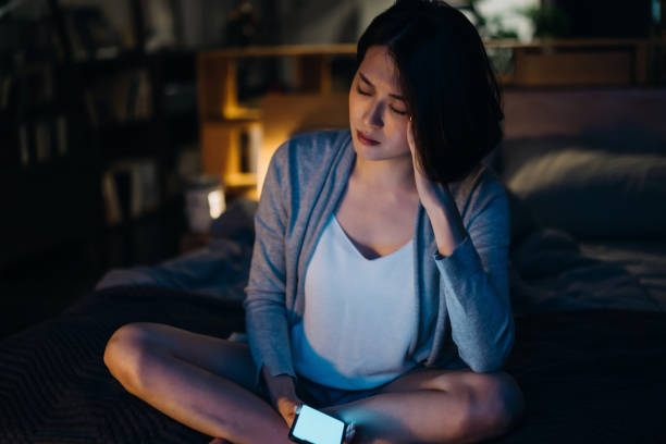 wanita muda asia stres dengan smartphone duduk di tempat tidur di kamar tidur dengan tangan di kepala, menghadapi masalah tidur dan insomnia di malam hari - resah potret stok, foto, & gambar bebas royalti