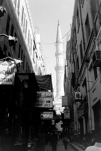 Cairo, Egypt - May 15, 1989: A narrow crowded alley between Bab Zuweila and  the famous Khan El Khalili bazaar in the historic centre of Islamic Cairo. Most of the people are wearing traditional clothes. In the Background are the minarets of a Mosque which are usually used  as orientation in this narrow surroundings. This black and white image was scanned from a negative.