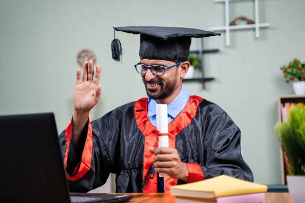 Student attending virtual graduation from laptop by holding certificate in graduation dress from home - concept of virtual celebrations, new normal lifestyle during coronavirus or covid-19 outbreak. Student attending virtual graduation from laptop by holding certificate in graduation dress from home - concept of virtual celebrations, new normal lifestyle during coronavirus or covid-19 outbreak online degree stock pictures, royalty-free photos & images