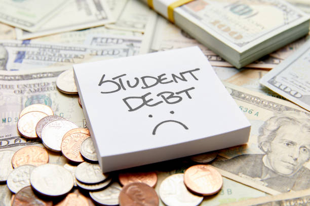 Student debt with sad face written on white sticky note on top of cash money with stack of money and coins stock photo