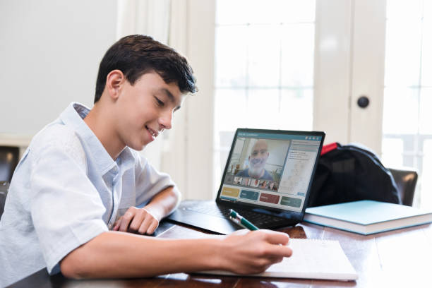 Student learns online during COVID-19 pandemic During a stay-at-home order, a middle schoolboy works on a homework assignment while distance learning. A male teacher is teaching via live stream. middle school teacher stock pictures, royalty-free photos & images