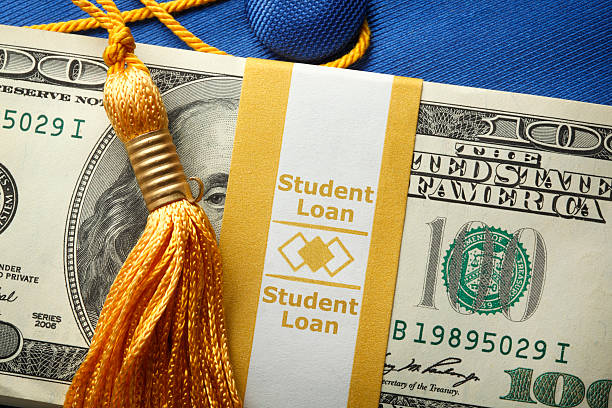 Student Loan Money On A Graduation Cap A stack of one hundred dollar bills in a money wrapper labeled "Student Loan" on top of a blue graduation cap.  A gold graduation tassel is draped over the stack of money. student loan stock pictures, royalty-free photos & images
