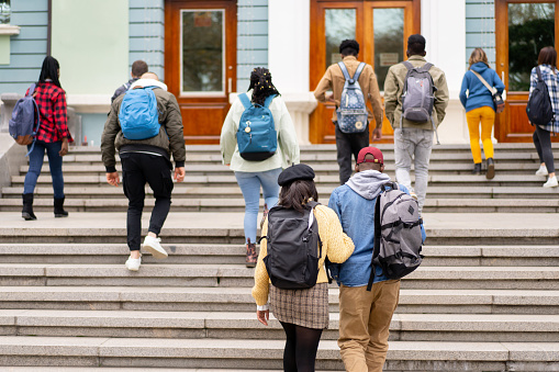 On a cold day the students are climbing up the steps towards the university entrance, everyone wearing coats and carrying backpacks containing their education books